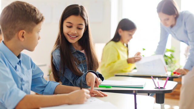 Positive European schoolgirl helping friend with task, pointing at boy's copybook, sitting near at desk in classroom of private school during lesson