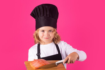 Kid cook hold cutting board with fish salmon steak and knife. Child chef cook. Child wearing cooker uniform and chef hat preparing food, studio portrait.