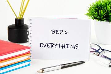A spiral notebook declares bed over everything. It sits alongside books, a pen, glasses, a plant, and a fragrance diffuser on a white surface, capturing a funny lifestyle concept