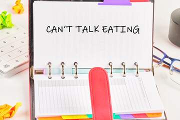 Open binder agenda shows the text cant talk eating printed on the top page. Nearby are crumpled papers, a calculator, eyeglasses, and other office items on a white surface