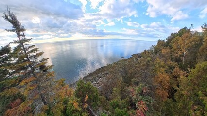 A panoramic view of the lower Saint Lawrence river reflecting a cloudy blue sky from a rocky coastal overlook bordered by autumnal evergreen and deciduous trees