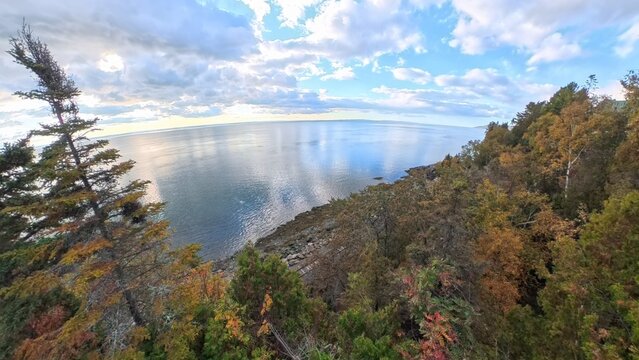A panoramic view of the lower Saint Lawrence river reflecting a cloudy blue sky from a rocky coastal overlook bordered by autumnal evergreen and deciduous trees