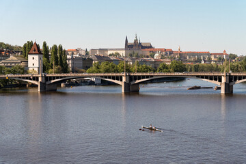Obraz premium Prague, Czech Republic - April 21, 2025: A peaceful scene featuring two rowers gliding across a Vlatva river beneath a wide stone bridge, with a backdrop of charming historic buildings and a castle