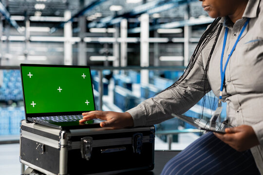 Mockup laptop next to female engineer in server hub implementing data backup and recovery solutions. African american data center employee using green screen notebook, optimizing network management