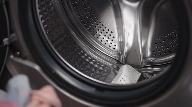 Man loading colorful pile of dirty clothes and placing detergent pod into open drum of modern front-loading washing machine, illustrating everyday housework and routine maintenance, close-up.