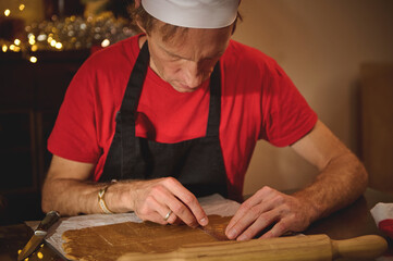Baker In Red Shirt And Black Apron Prepares Dough, Cutting Gingerbread Sheets Under Warm Christmas Lights