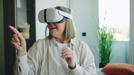 Senior woman exploring virtual reality inside her modern home while using VR headset during a sunny afternoon - Powered by Adobe
