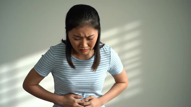 Young woman in a striped shirt clutches her stomach looking distressed against a plain wall with faint window-blind shadows
