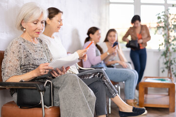Naklejka na ściany i meble Interested senior woman sitting in lobby of clinic with papers in hand, reading medical assessment report while waiting for appointment with doctor