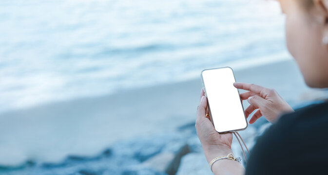 Person using smartphone with blank white screen at seaside,Asian Traveler siting by sea beach typing message concept representing travel communication,social media connection,modern digital lifestyle