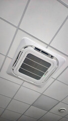 Ceiling-mounted cassette air conditioner unit with a smoke detector on a white suspended ceiling in a modern office environment