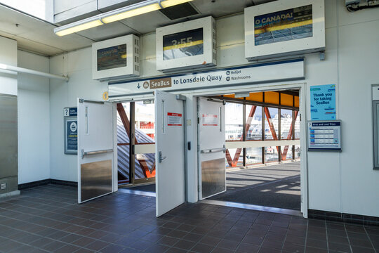 Open entrance doors at Vancouver SeaBus terminal to Lonsdale Quay, featuring digital clocks, schedules, Helijet signs, and harbor views