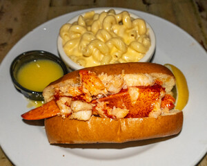 A classic lobster roll, generously filled with succulent lobster meat, served on a toasted bun alongside a side of creamy macaroni and cheese, melted butter, and a lemon wedge.