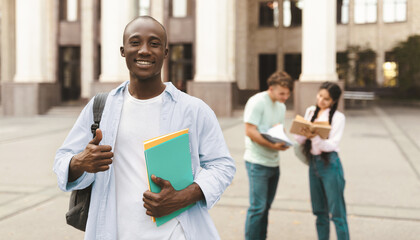 Happy african american student guy standing outdoors with his classmates in university campus, posing and smiling to camera. Study after covid-19 quarantine concept