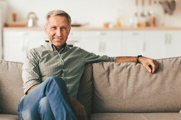 Fototapeta premium Contented Senior Man Sitting On Sofa At Home, Smiling Looking At Camera. Retired Male Relaxing On Couch In Modern Living Room Indoor On Weekend. Retirement Lifestyle, Domestic Comfort Concept