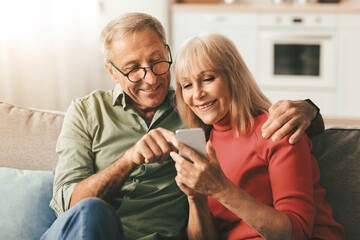 Fototapeta na wymiar Happy Senior Couple Using Mobile Phone Together, Husband Pointing Finger At Cellphone Showing Something Browsing Internet Sitting On Couch At Home. Mature People And Gadgets