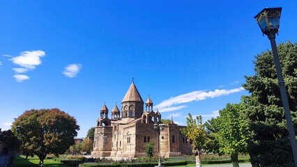 A historic stone church with conical domes, surrounded by lush gardens and trees under a vibrant...
