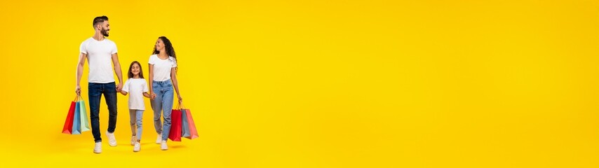 Fototapeta premium Family Shopping. Middle-Eastern Parents And Daughter Carrying Colorful Shopper Bags Walking Together Holding Hands Posing Over Yellow Background In Studio. Full Length