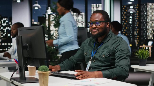 Black male consultant focused on IT company development with digital tools, review analytics and performance metrics at workstation. Staff planning solutions and aligning objectives. Camera B.