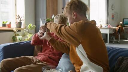Medium shot of young teenage boy fighting with brother for TV remote while sitting on couch together and eating popcorn - Powered by Adobe