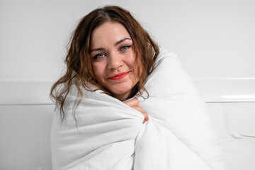 Young woman with wavy brown hair wrapped snugly in a white blanket sits indoors and smiles softly at camera against minimal white wall and bedding creating a cozy warm mood and relaxed morning comfort