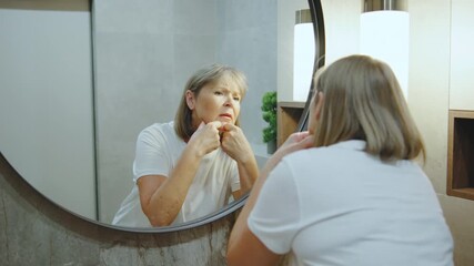 Senior woman performing self-care routine in a bright bathroom while examining her skin in a round mirror during the afternoon - Powered by Adobe