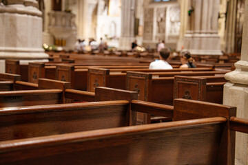 Wooden church pews fill the foreground, leading to a softly lit cathedral interior. A few people are seated, creating a quiet and reverent atmosphere in the sacred space.