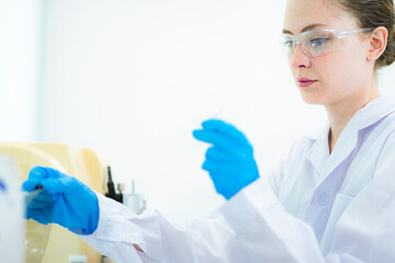 In a bright modern lab, a dedicated female scientist holds a glass slide to the light. She carefully observes the results of her experiment with focused curiosity and a hopeful expression.