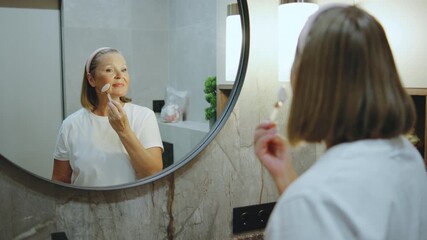 Elderly woman applying skincare in a modern bathroom while smiling at her reflection in the round mirror - Powered by Adobe