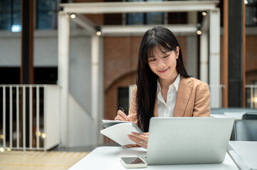 Asian businesswoman or office worker writing in notebook while looking at laptop on table in a cafe.