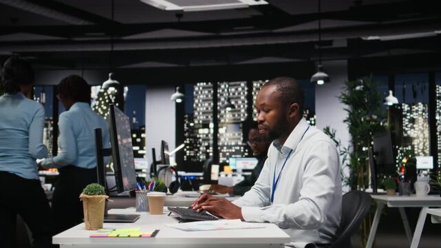 Black business consultant proofreading financial numbers at desktop after hours, studying data for monthly performance reports. Ensuring company objectives achievement at night. Camera A.