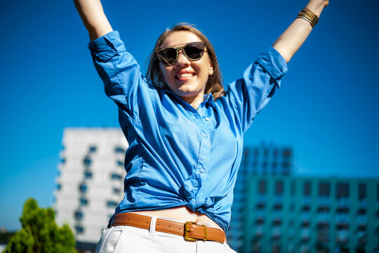 Fototapeta Young woman with blonde hair smiles brightly while celebrating outdoors in a blue shirt, surrounded by modern buildings under a clear sky.