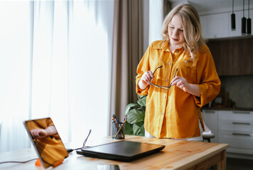 happy blonde business woman stands in home office, adjusting glasses and focused on laptop