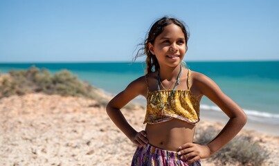 a cute Indigenous Australian girl aged 11 years old, standing with her hands on her hips looking proud. She's standing on a beach on a beautiful sunny day
