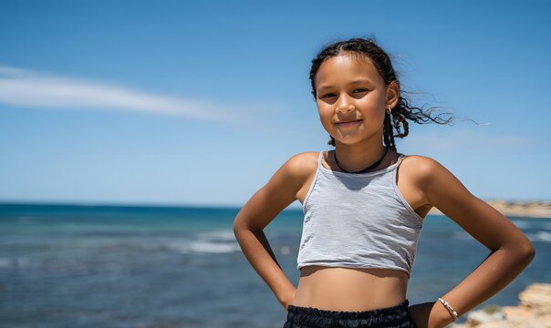a cute Indigenous Australian girl aged 11 years old, standing with her hands on her hips looking proud. She's standing on a beach on a beautiful sunny day