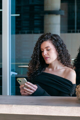 Young Argentinian woman with curly hair looking at her smartphone in a modern urban café. Natural light and neutral expression.