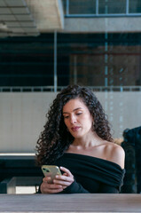 Young woman with curly hair using her phone while sitting at an outdoor café. Neutral expression, casual and modern style.