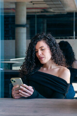 Adult Argentinian woman with curly hair using her phone in a stylish outdoor restaurant. Calm and natural look.