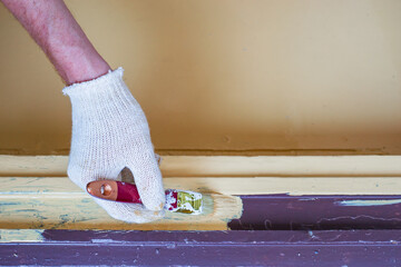 person wearing protective glove is applying paint to wooden surface using brush. setting appears to be home renovation, featuring bright wall colors. close up.