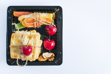 black tray holds an appealing selection snacks. there are three bright red cherries, crunchy crackers, dried fruits, and nuts, all tied together with twine. close up.