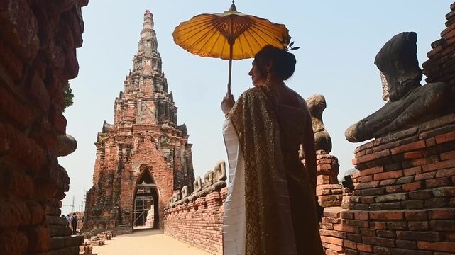 Ayutthaya, Thailand &ndash; March 2023: Woman in traditional Thai dress holding yellow umbrella walking by headless Buddha statues at Wat Chaiwatthanaram, UNESCO World Heritage Site symbol of ancient Siam.