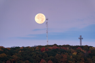 A supermoon rises above autumn treetops and a cross, with a radio tower silhouette. g.