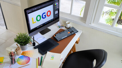 Top view of graphic designer desk with computer screen showing colorful logo design, drawing...