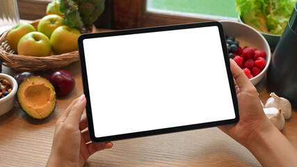 Hands holding digital tablet with blank screen above wooden table filled with fresh fruits. Health and nutrition concept