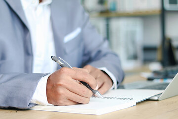 Businessman writing in notebook with pen while working at office desk with laptop. Concept of business planning, office work, productivity, and corporate management.