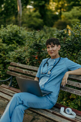 Female medical worker in scrubs using a laptop on a park bench