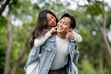Young asian couple fooling around woman poking man's cheeks while getting piggyback walking in park.