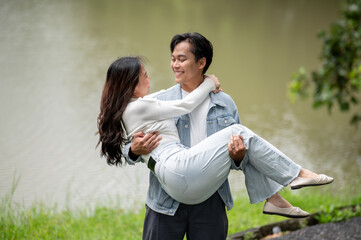 Young asian couple fooling around as a man princess carrying woman standing walking outdoors in park
