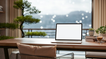 Blank screen laptop with books and coffee on wooden table across bonsai tree and snow outside balcony.