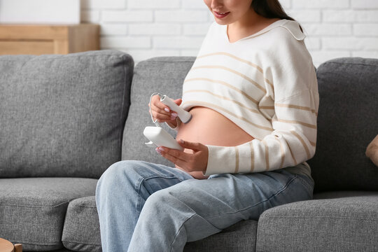 Young pregnant woman with fetal doppler listening to baby's heartbeat on sofa at home